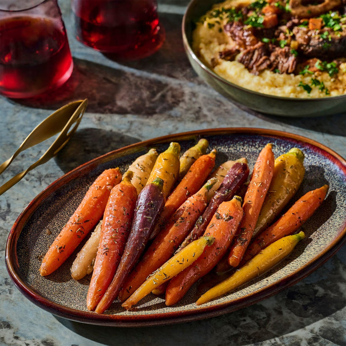 A plate of colorful roasted carrots sits alongside a bowl of braised meat over polenta and two glasses of red wine, suggesting a gourmet meal.