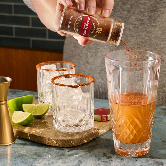 A person is adding Tajin seasoning to a cocktail, with other cocktails and lime wedges nearby.