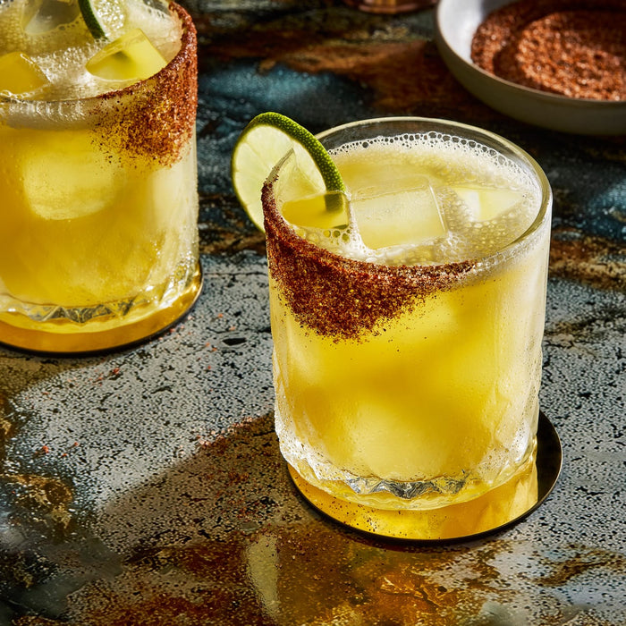 Two vibrant cocktails with chili-lime rims and lime wedges sit on a textured surface, suggesting a refreshing and spicy beverage.