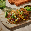 Two appetizing tacos topped with pineapple salsa and lime wedges are presented on a marble board.