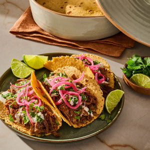 A plate of delicious tacos topped with pickled onions and cilantro, accompanied by lime wedges and a bowl of tortillas.