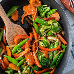 A close-up of a wok filled with stir-fried vegetables, including carrots, broccoli, peppers, and snow peas.