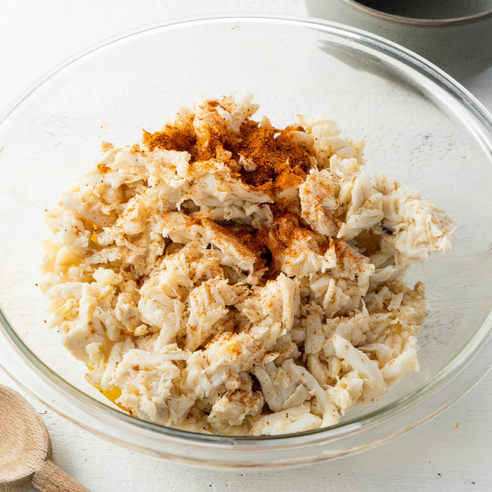 A hand sprinkles Old Bay seasoning on a bowl of crab meat.