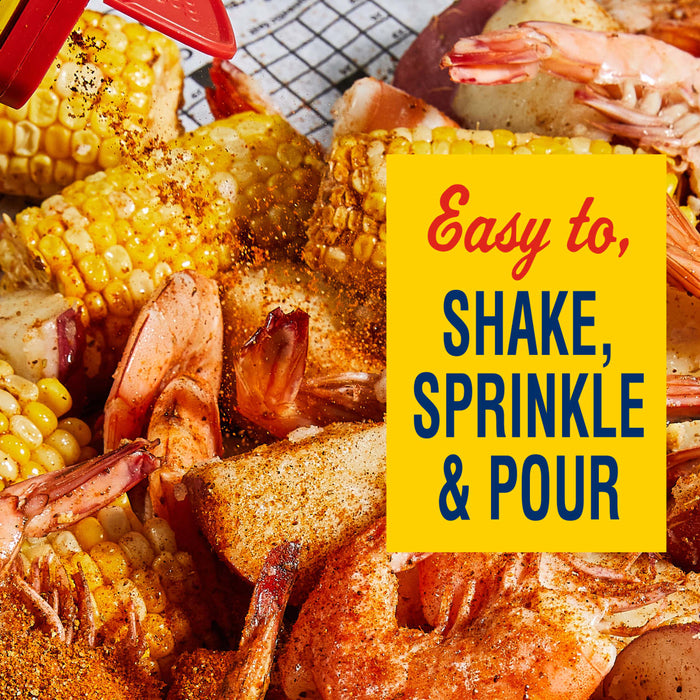 A hand is pouring Old Bay seasoning over a bowl of shrimp, corn, and potatoes.