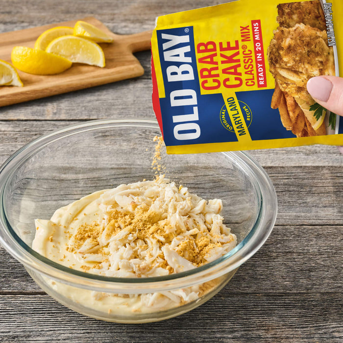 A person pours a packet of Old Bay Regular Crab Cake Seasoning Mix 1.24 Oz into a glass bowl containing fresh lump crab meat and creamy mayonnaise on a rustic wooden table.