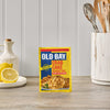 A package of Old Bay Regular Crab Cake Seasoning Mix 1.24 Oz stands on a wooden countertop in a bright kitchen setting next to fresh lemons and cooking utensils.