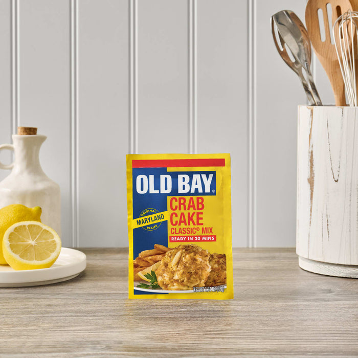 A package of Old Bay Regular Crab Cake Seasoning Mix 1.24 Oz stands on a wooden countertop in a bright kitchen setting next to fresh lemons and cooking utensils.