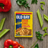 A yellow packet of Old Bay Regular Crab Cake Seasoning Mix 1.24 Oz is centered on a rustic wooden surface surrounded by fresh red bell peppers, celery stalks, and sprigs of parsley.