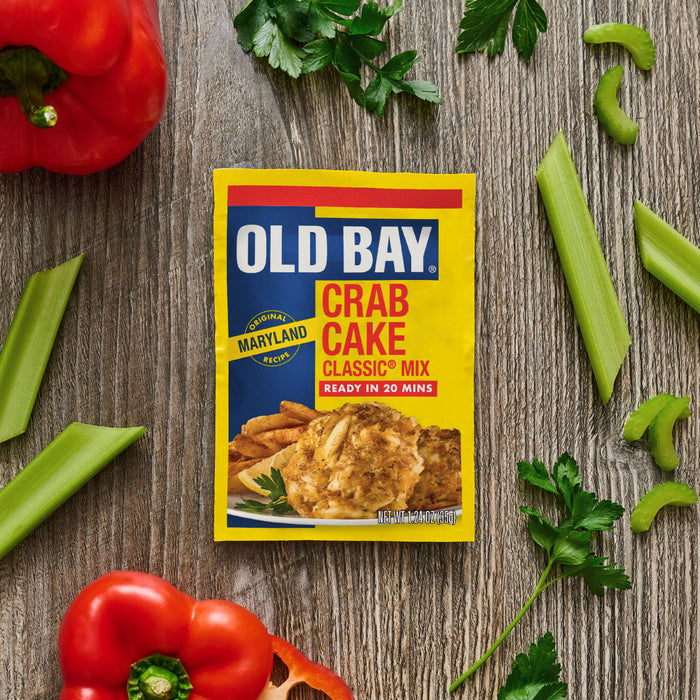 A yellow packet of Old Bay Regular Crab Cake Seasoning Mix 1.24 Oz is centered on a rustic wooden surface surrounded by fresh red bell peppers, celery stalks, and sprigs of parsley.