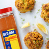 A bottle of Old Bay Seasoning next to four crab cakes with lemon wedges and green onions.