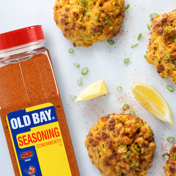 A bottle of Old Bay Seasoning next to four crab cakes with lemon wedges and green onions.
