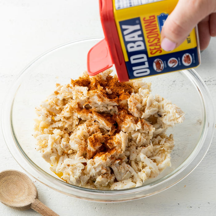 A hand sprinkles Old Bay seasoning on a bowl of crab meat.
