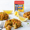 A container of Old Bay Seasoning next to crab cakes and lemon wedges.