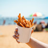 A hand holds a white cup filled with french fries on a beach.