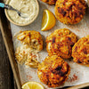 A tray of golden-brown crab cakes is served with a creamy dipping sauce and lemon wedges.