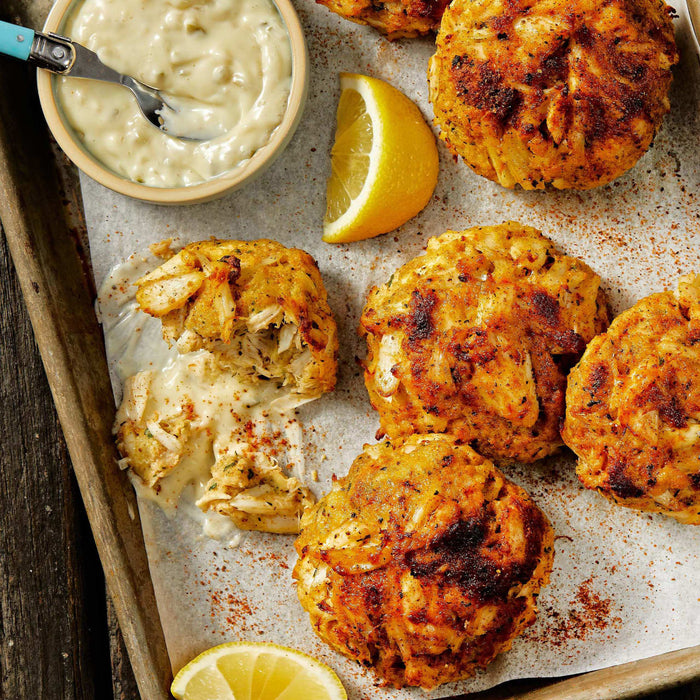 A tray of golden-brown crab cakes is served with a creamy dipping sauce and lemon wedges.