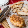 A seasoned chicken breast is displayed on parchment paper, partially sliced, alongside serving tongs.