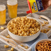 A hand sprinkles Old Bay Black Bird Seasoning, 6.5 Oz from a yellow and blue tin onto a large bowl of popcorn served on a wooden tray with pretzels and two glasses of beer.