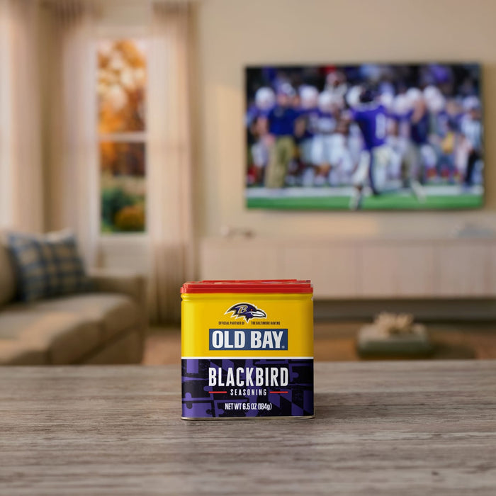 A tin of Old Bay Black Bird Seasoning, 6.5 Oz sits on a wooden table in a living room with a football game playing on the television in the background.