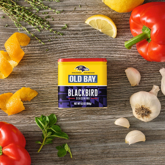 A tin of Old Bay Black Bird Seasoning, 6.5 Oz is centered on a rustic wooden surface surrounded by fresh ingredients like red bell peppers, garlic cloves, lemon slices, orange peels, and sprigs of thyme and oregano.