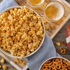 A large bowl of popcorn is generously coated with Old Bay Black Bird Seasoning, 6.5 Oz and served alongside pretzels and glasses of beer for a game day snack.
