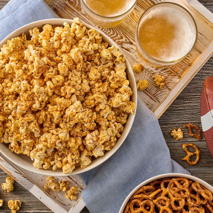 A large bowl of popcorn is generously coated with Old Bay Black Bird Seasoning, 6.5 Oz and served alongside pretzels and glasses of beer for a game day snack.