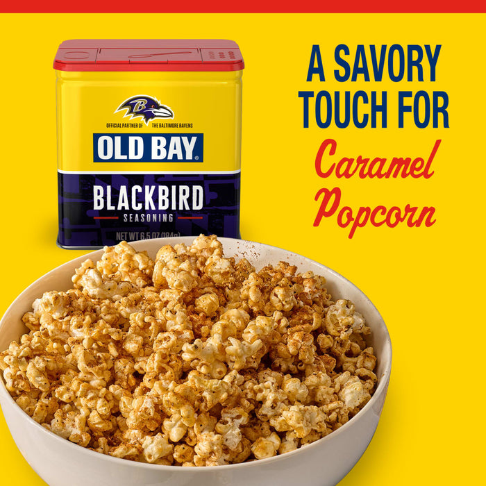 A bowl of caramel popcorn is dusted with Old Bay Black Bird Seasoning, 6.5 Oz next to the yellow and purple Baltimore Ravens themed tin.