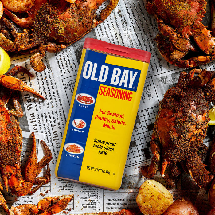 A can of Old Bay seasoning sits on top of a newspaper with crab legs and potatoes.