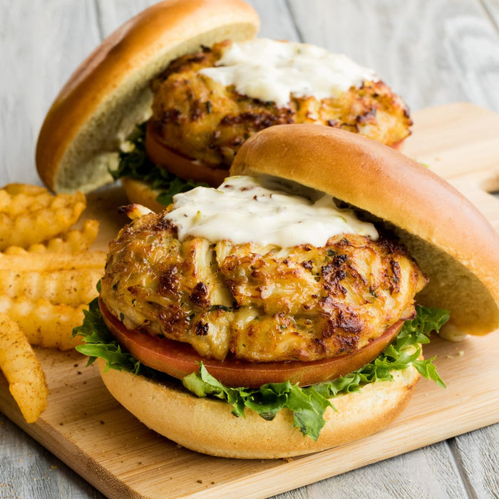 Two crab cake sandwiches with lettuce, tomato, and a creamy sauce on a wooden cutting board with french fries.
