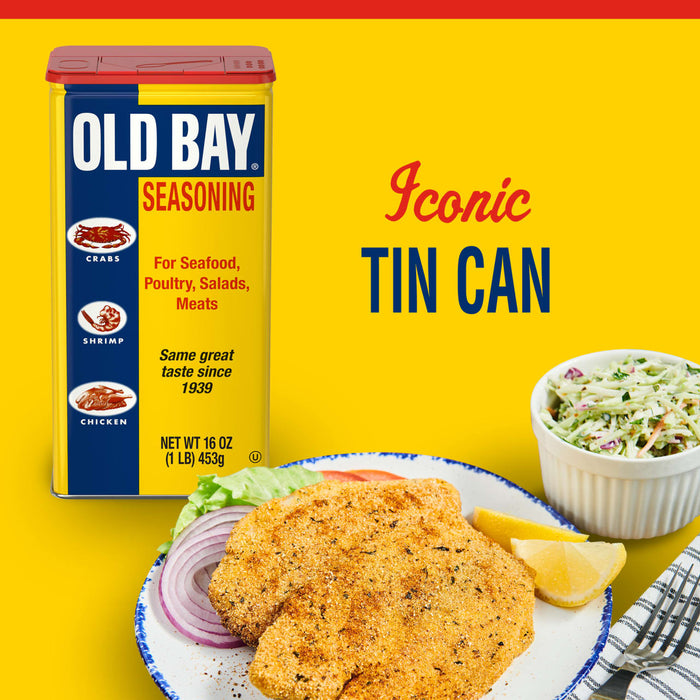 A container of Old Bay Seasoning next to a plate of fried fish, lemon wedges, and red onion with a bowl of coleslaw and a fork.