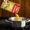 A box of Zatarain's Yellow Rice Mix is being poured into a pot on a stove.