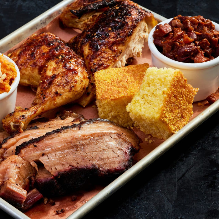 A tray of barbecue featuring brisket, chicken, cornbread, baked beans, and sweet potatoes.