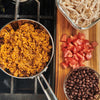 A close-up of a pot of rice, a bowl of black beans, chopped tomatoes, and shredded chicken.