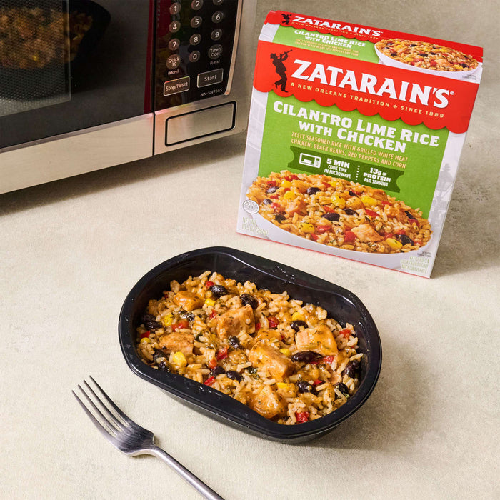 A microwave, a box of Zatarain's Cilantro Lime Rice with Chicken, and a cooked meal in a black container with a fork are on a table.