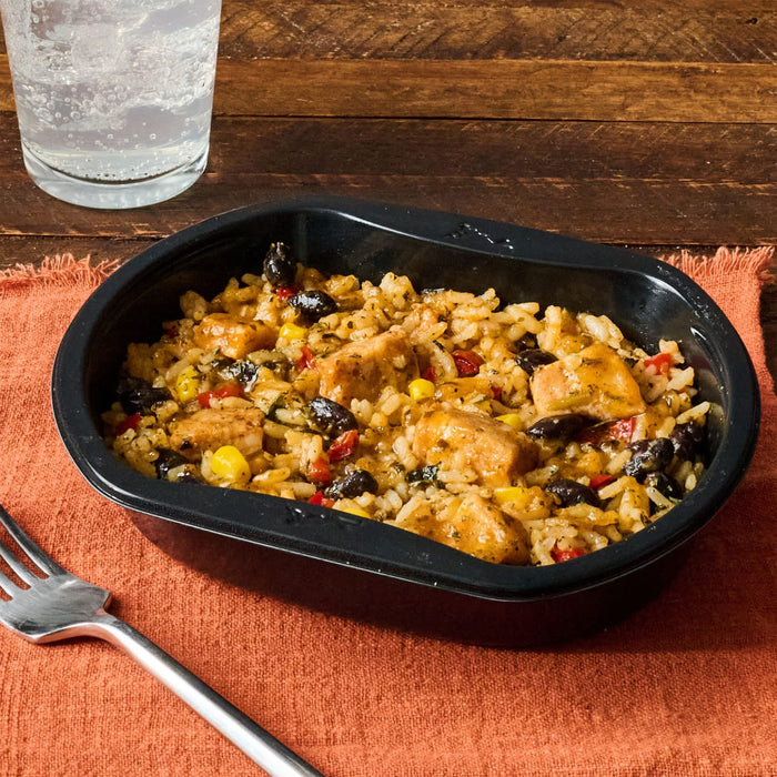 A black tray filled with a rice and chicken dish sits on an orange cloth next to a glass of sparkling water and a fork.