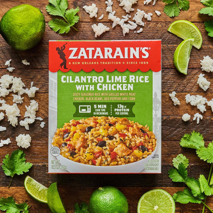 A box of Zatarain's Cilantro Lime Rice with Chicken is surrounded by limes, cilantro, and cooked rice on a wooden surface.