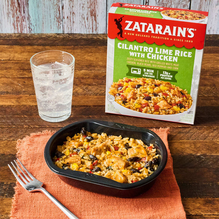 A box of Zatarain's Cilantro Lime Rice with Chicken is displayed next to a cooked meal and a glass of sparkling water on a wooden table.