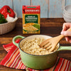 A person stirs a pot of cooked rice, with a box of Zatarain'S Garlic Parmesan Long Grain White Rice, 6.9 Oz, sitting on the table behind it.