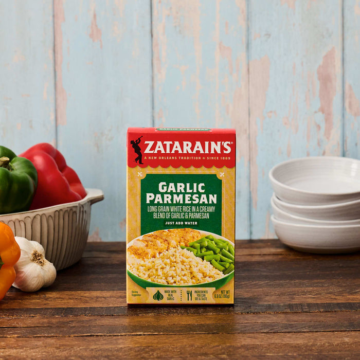 A box of Zatarain'S Garlic Parmesan Long Grain White Rice, 6.9 Oz, is displayed on a rustic wooden table surrounded by fresh ingredients like bell peppers and garlic.