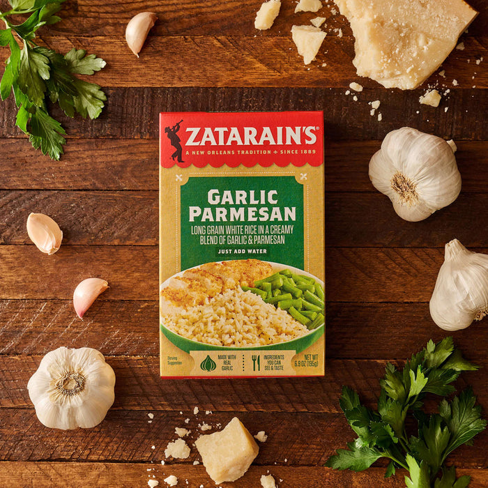 A box of Zatarain'S Garlic Parmesan Long Grain White Rice, 6.9 Oz, is displayed on a wooden surface, surrounded by its fresh ingredients, including whole garlic bulbs, cloves, a wedge of Parmesan cheese, and fresh parsley.