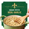A hand stirs a pot of cooked Zatarain'S Garlic Parmesan Long Grain White Rice, 6.9 Oz with a wooden spoon, shown in front of a green label that reads Made With Real Garlic.