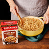 A person is holding a yellow pot of Zatarain's Dirty Rice with a wooden spoon.