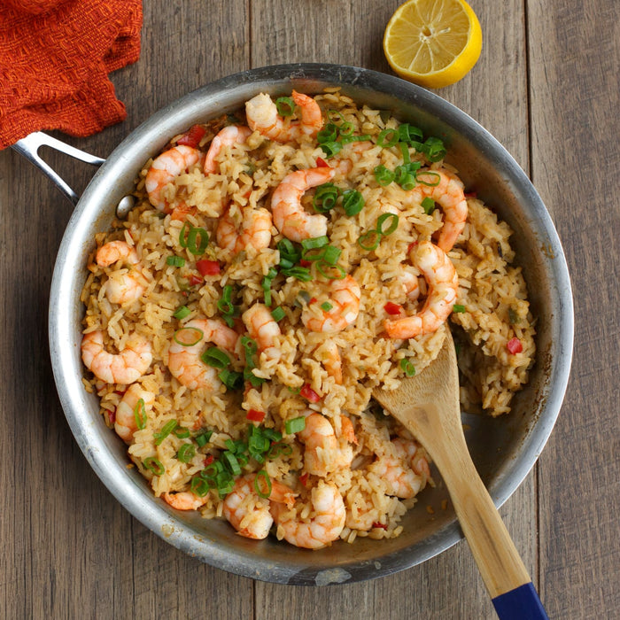 A pan of shrimp and rice dish with a wooden spoon and a lemon on a wooden table.