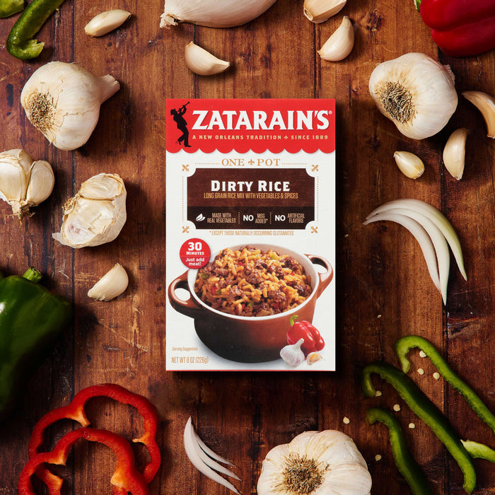 A box of Zatarains Dirty Rice mix is surrounded by garlic peppers and onions on a wooden surface.
