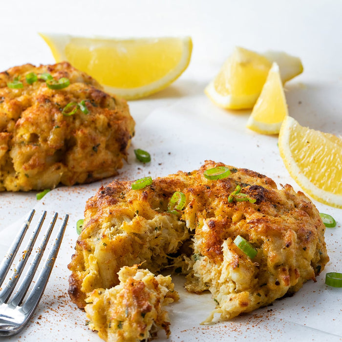 Two golden-brown crab cakes, garnished with green onions and served with lemon wedges, are displayed on a white surface.