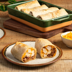 A plate with two chicken burritos, one cut in half to show the filling, sits in front of a baking dish filled with more burritos and a bowl of shredded cheese.