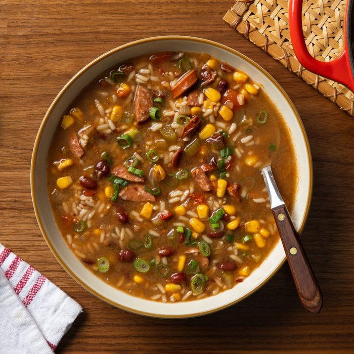 Overhead shot of a bowl of hearty soup with corn, beans, rice, and sausage, garnished with green onions, and a spoon resting inside.