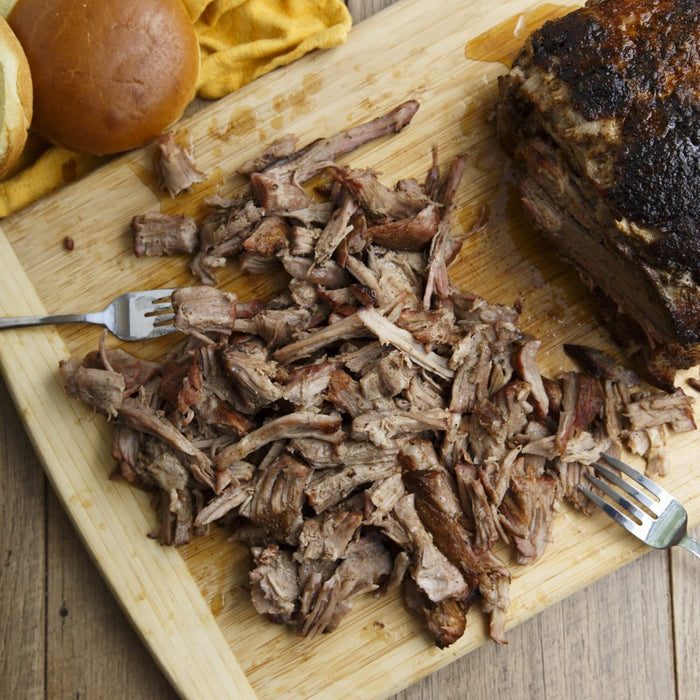 A wooden cutting board displays pulled pork, a whole piece of pork, two forks, and buns, suggesting a meal preparation or serving.