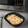 A black plastic container of shrimp alfredo pasta sits on a gray countertop next to a stainless steel microwave.