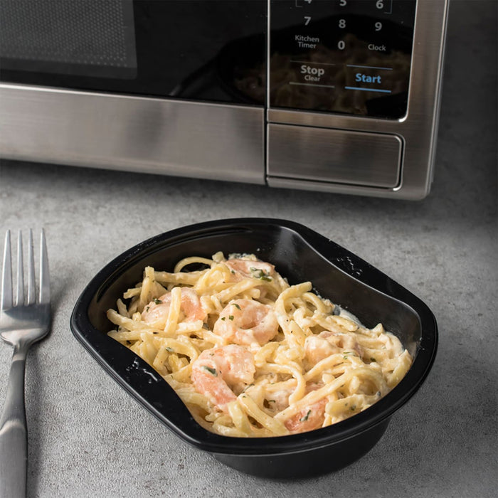 A black plastic container of shrimp alfredo pasta sits on a gray countertop next to a stainless steel microwave.